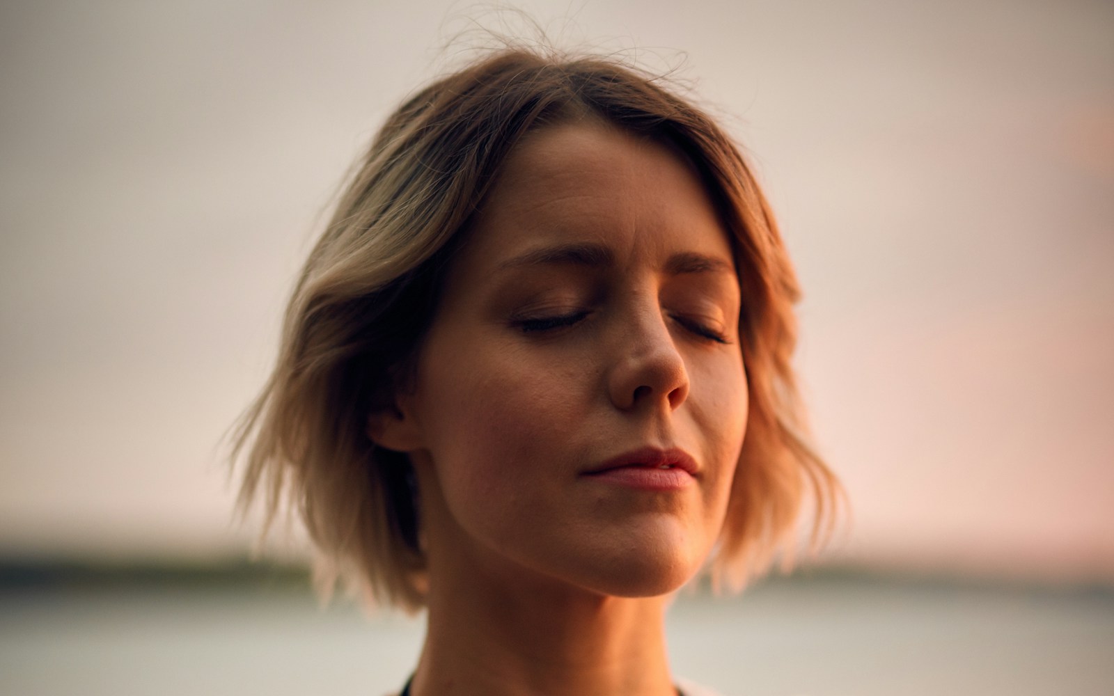 Femme aux yeux fermés respirant calmement au bord de l'eau, le visage baigné d'une lumière dorée de fin de journée.